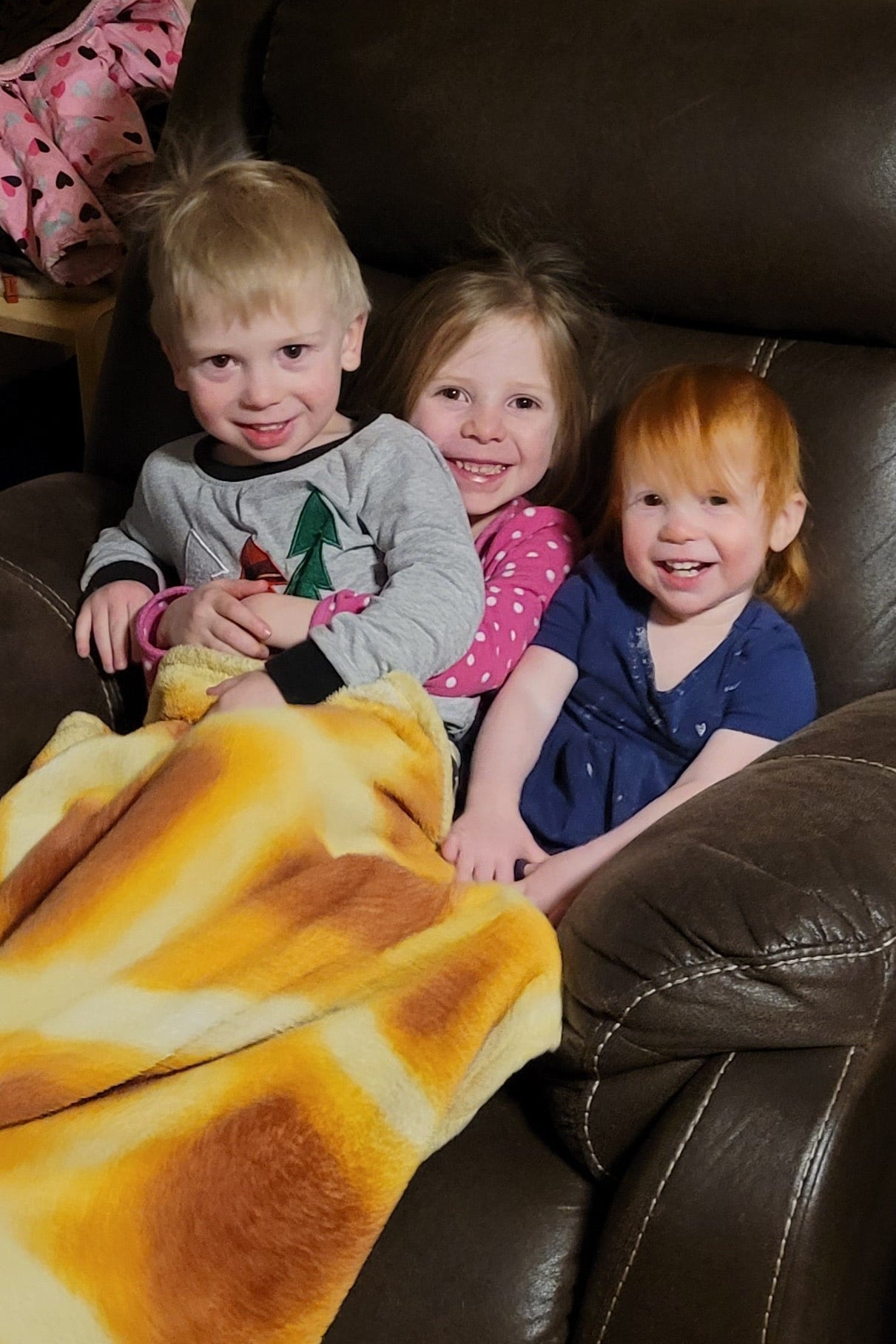 Three children lying on a brown leather couch with a yellow blanket