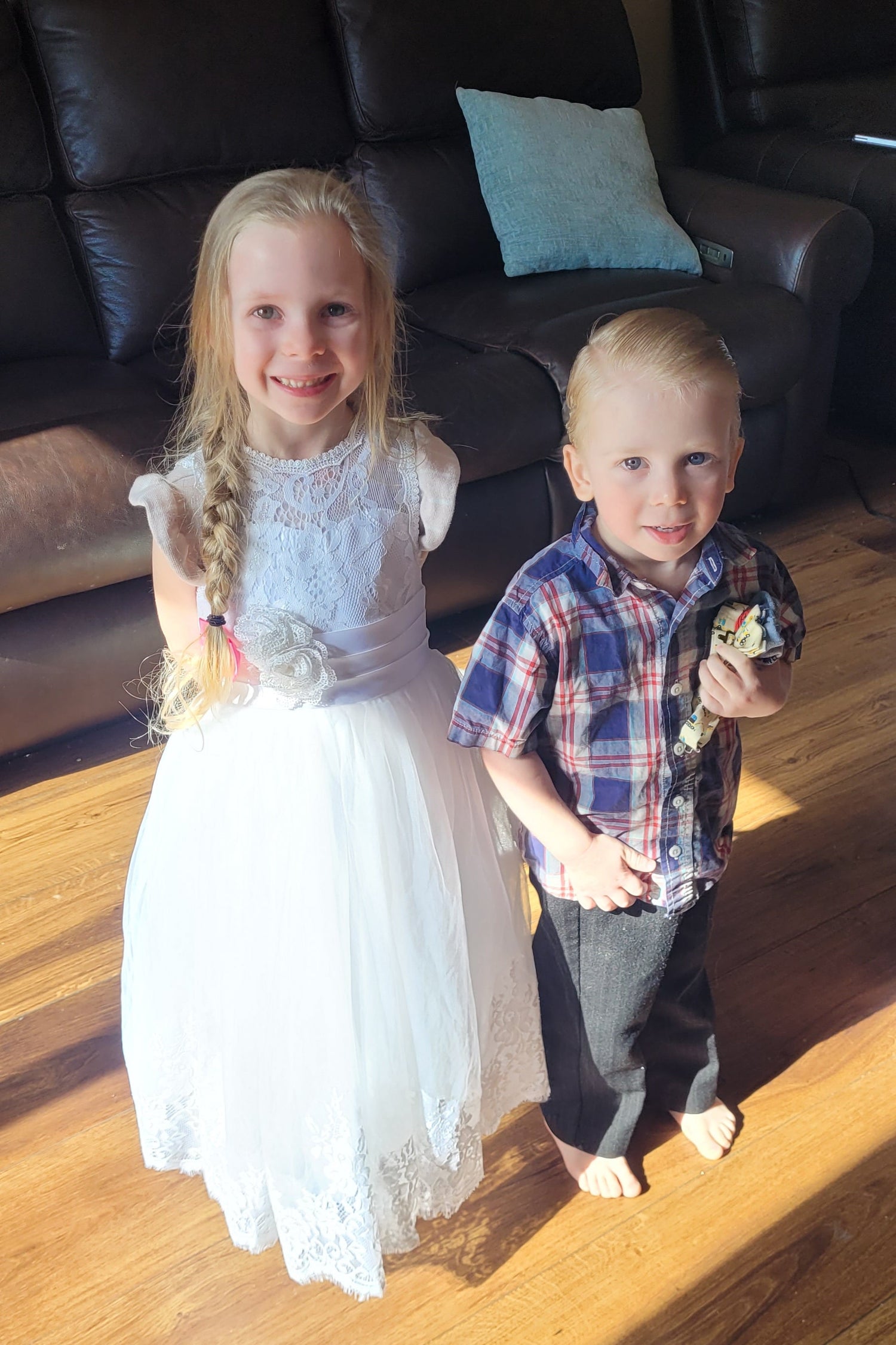 Two children, a boy and a girl, standing on a wooden floor with a couch in the background.