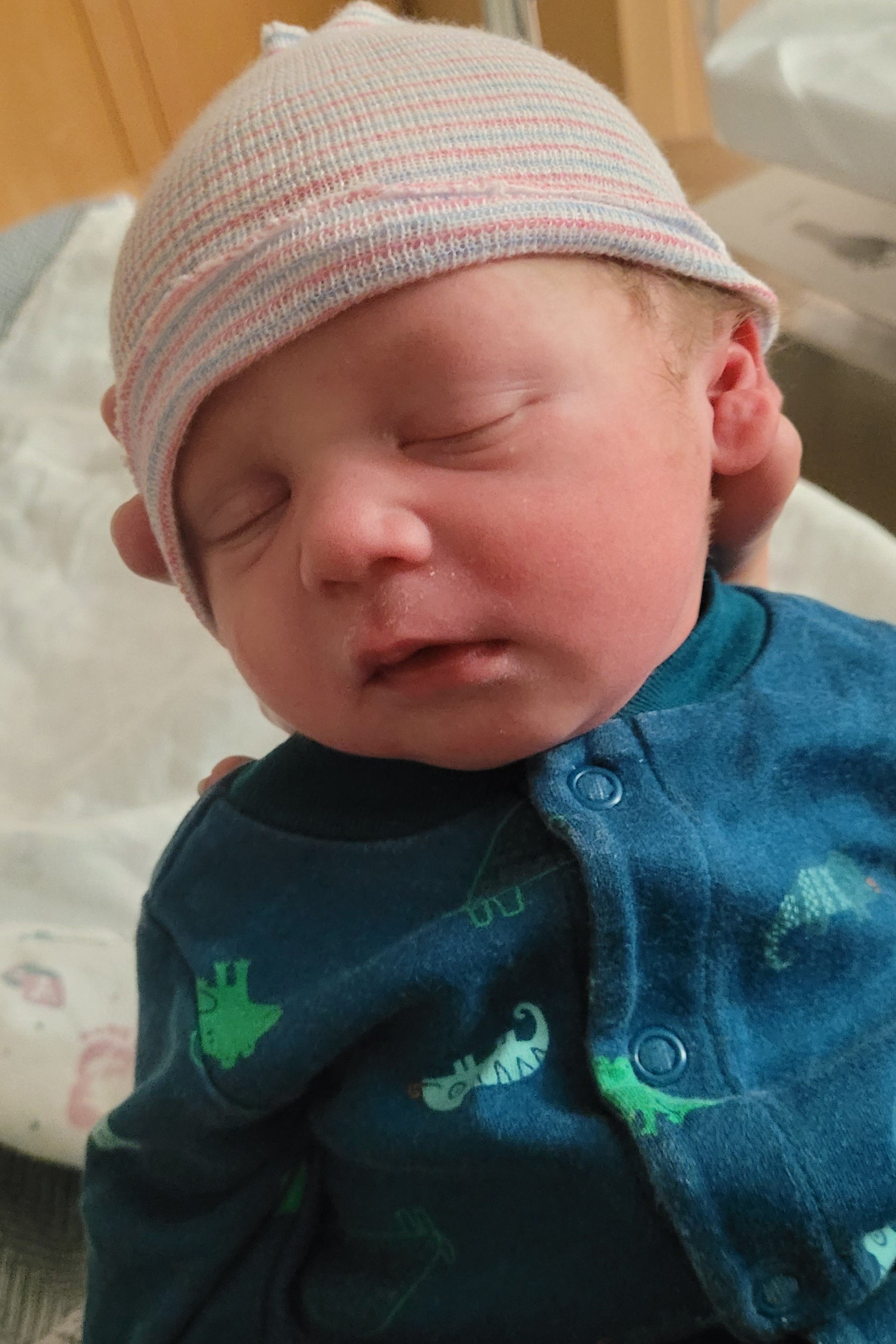 Newborn baby wearing a pink hat and green patterned outfit in a hospital setting.