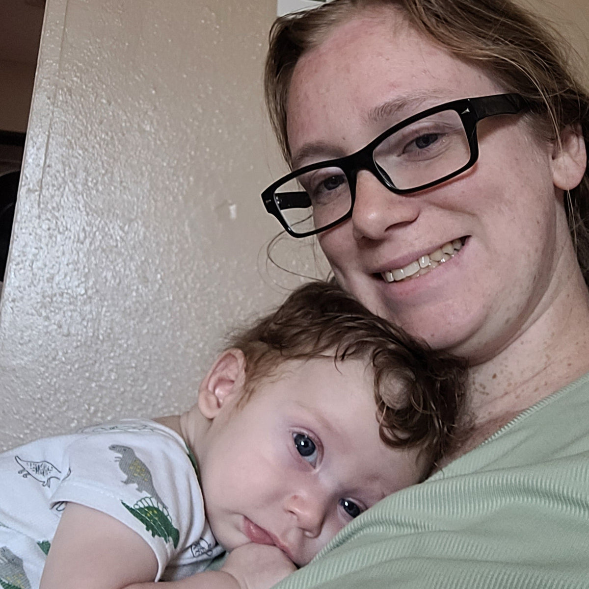 A woman wearing black-rimmed glasses and a sage green shirt holds a baby in a home setting. A framed sign on the wall behind them reads 'I STILL FALL FOR YOU everyday'.